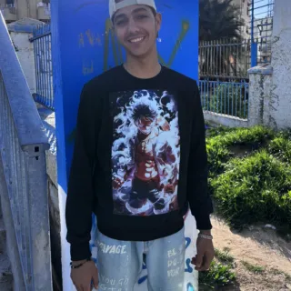 Un jeune homme souriant porte un sweatshirt noir en Réalité Augmentée avec un design animé. Il se tient en extérieur sous un beau ciel ensoleillé, appuyé contre un mur bleu décoré de graffitis. Son style décontracté avec une casquette à l’envers et un jean délavé ajoute une touche urbaine et tendance.