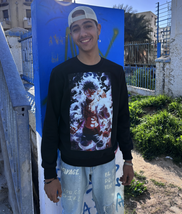 Un jeune homme souriant porte un sweatshirt noir en Réalité Augmentée avec un design animé. Il se tient en extérieur sous un beau ciel ensoleillé, appuyé contre un mur bleu décoré de graffitis. Son style décontracté avec une casquette à l’envers et un jean délavé ajoute une touche urbaine et tendance.